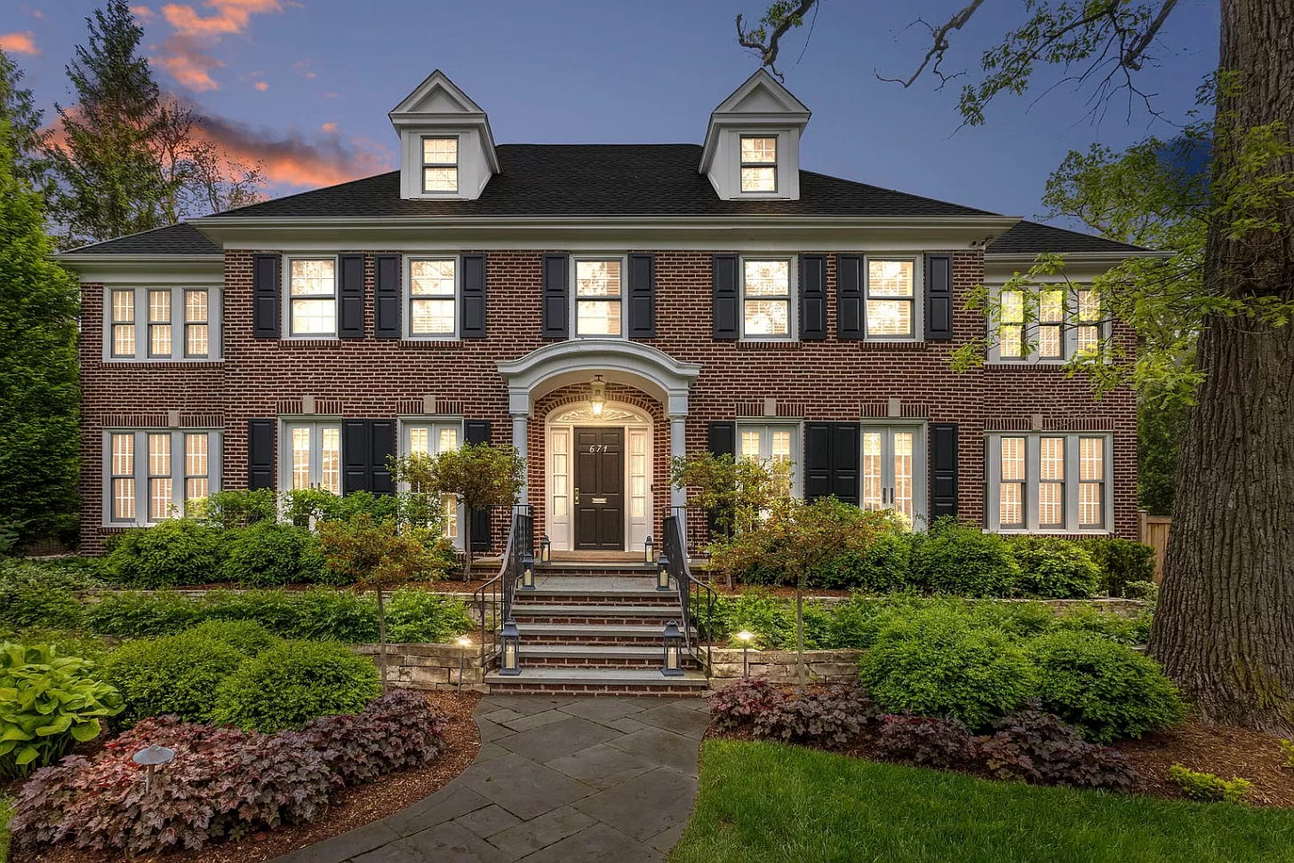 The front of the "Home Alone" house. It's a large brick house with 5 windows across the top, and 4 on the bottom (two on either side of the door). There are also two side additions to the house with two windows each, one on top and one on the bottom. The brick is red and the roof and shutters on the windows are black. The lawn has a lot of plants in it and 3 different levels alongside stairs up to the door. There are large trees on the side of the image, and the sun is setting in the background lighting up some clouds behind the house in a really pretty pink color.