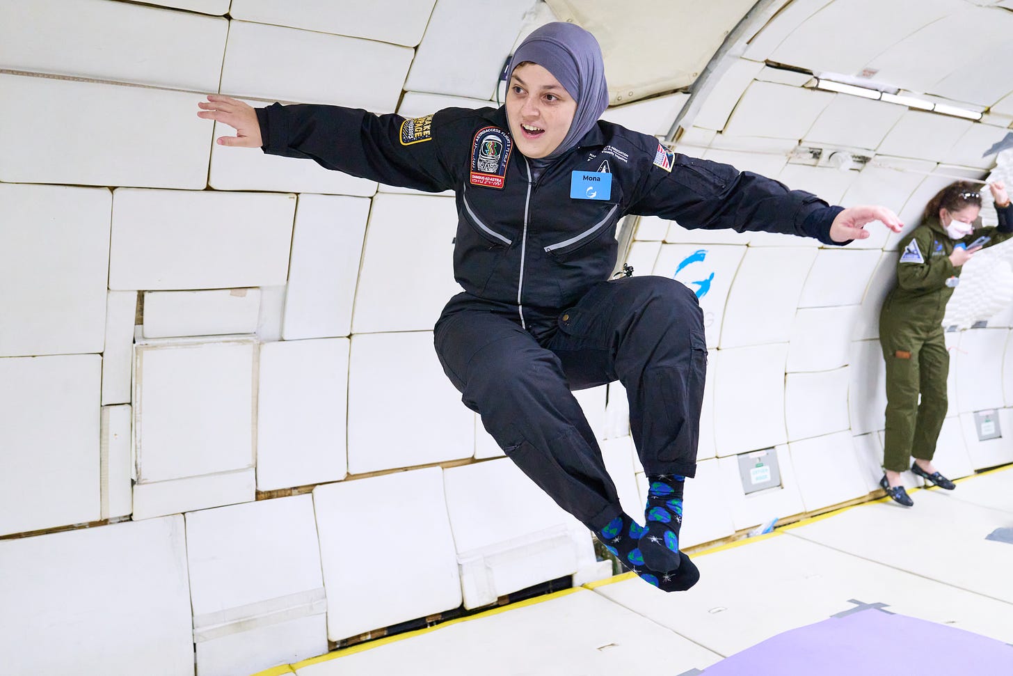 Dr. Mona Minkara is wearing a black flight jump suit and grey hijab. She is inside a G-Force One, a modified Boeing 727-200. Mona is smiling and has both hands outstretched wide, with her right hand on the side of the Zero-G aircraft. Both legs are bent at the knees as though Mona were sitting in a chair with her feet crossed. She is wearing a dark blue sock with lighter blue clouds on them and black Mary Jane style shoes as she floats weightlessly in the seatless  aircraft. In the background, is a dark haired woman in an olive green flight suit deck shoes. She’s wearing a mask and looking at her cell phone as she holds on to a bar on the side of the aircraft with her other hand.