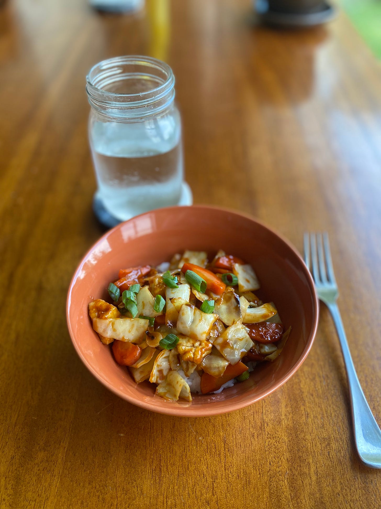 An orange bowl of white rice topped with the vegetable and tofu teriyaki described above. On a coaster behind it is a glass of water.