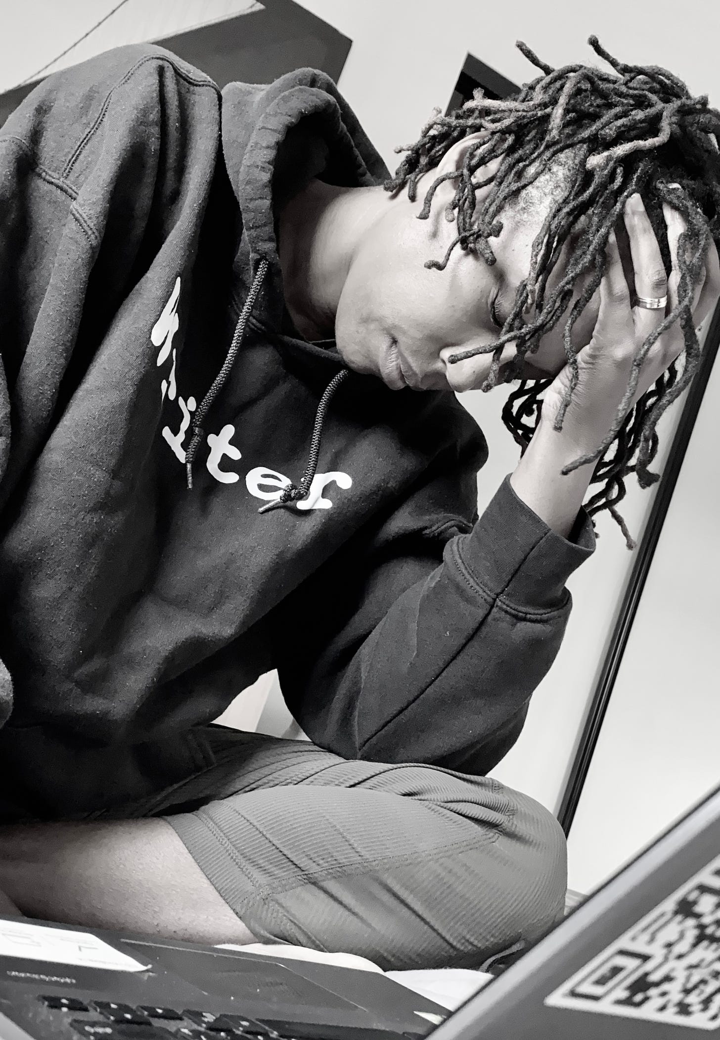 Sandra is a Black femme with locs, wearing a black hoodie with the word writer on it. The image is in black and white. Their head is in their hands looking at a lap top.