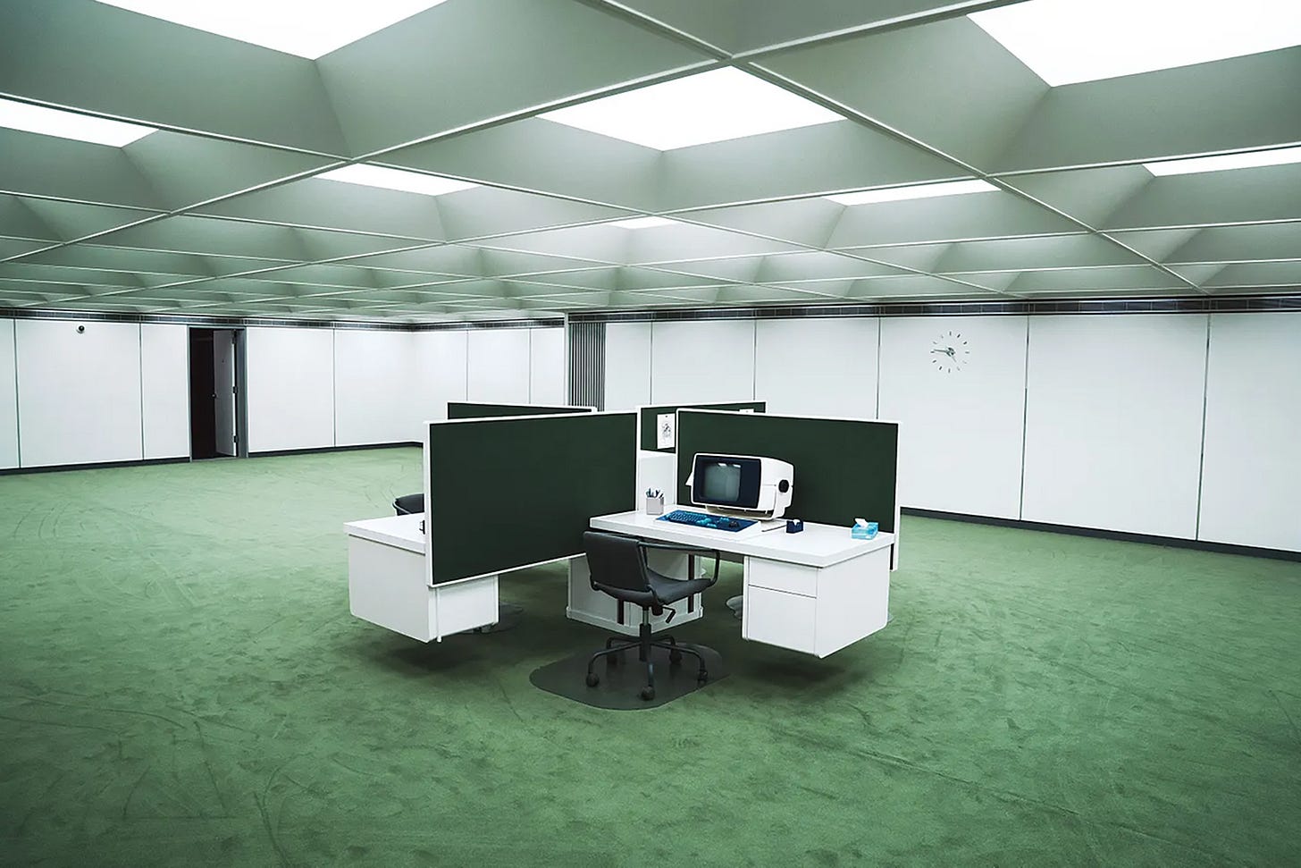 Photo of a low-ceiled office with a pale green carpet and four white desks arranged in the middle of the sparse room. 