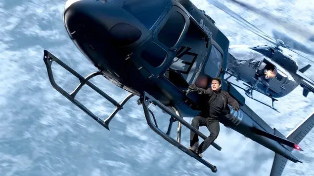 Tom Cruise as Ethan Hunt hanging off a helicopter against mountains. Tom Cruise as Ethan Hunt hanging off a helicopter against mountains.