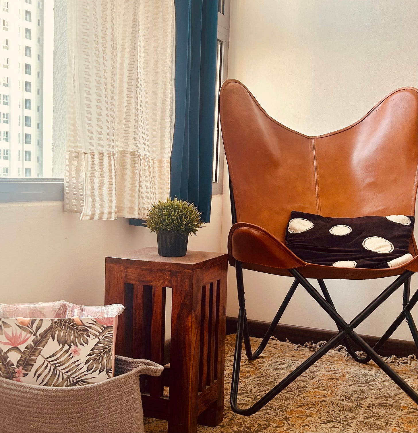 A cozy reading nook beside a window is decorated with a dhurrie, a leather chair,  a small table with a plant pot kept on it and a woven basket placed on the floor. 