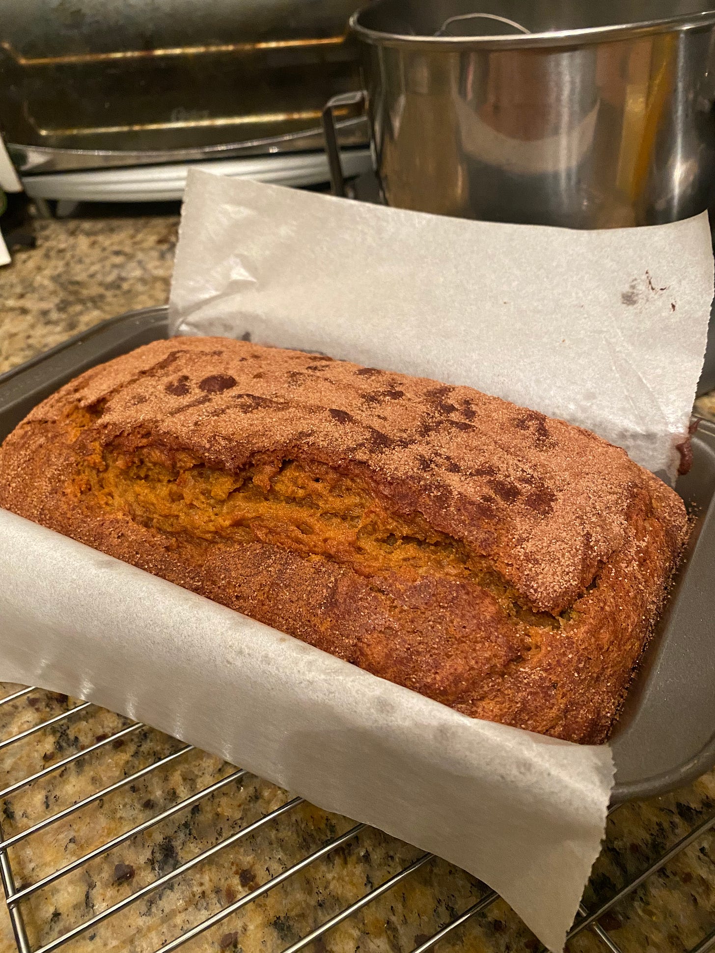 A loaf pan lined with parchment and filled with a cracked, sugar-crusted pumpkin loaf, resting on a cooling rack.