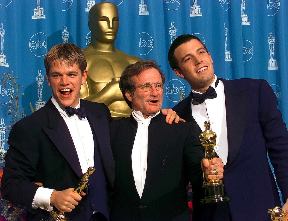 Matt Damon, Robin Williams, and Ben Affleck stand holding Academy Awards in front of a blue curtain and an oversized golden award statue