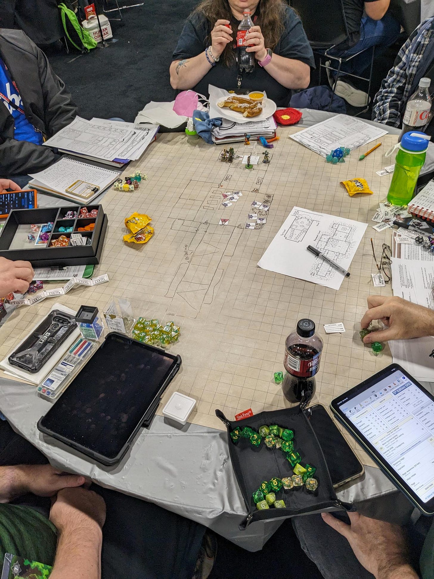 A gaming table with a dry erase map showing a dungeon and a battle in progress. Around the outside of the map sit players, half in frame, with snacks, dice, dice trays, and character sheets.