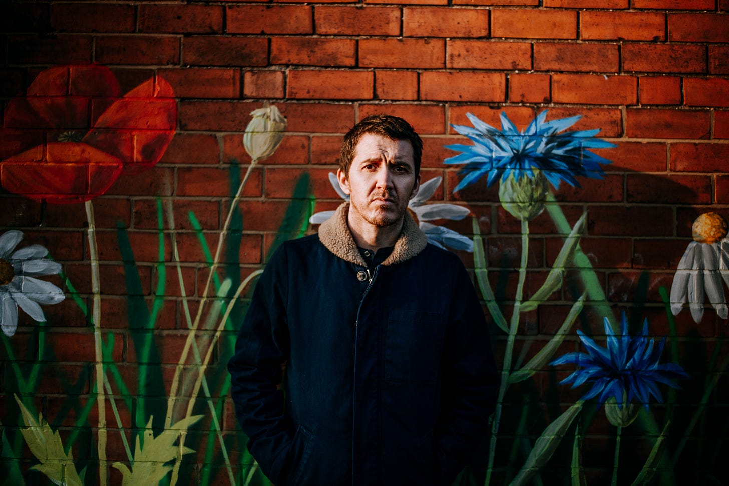 Neil Thomas songwriter and lead singer of Pavey Ark standing in front of a brick wall with flowers painted on it