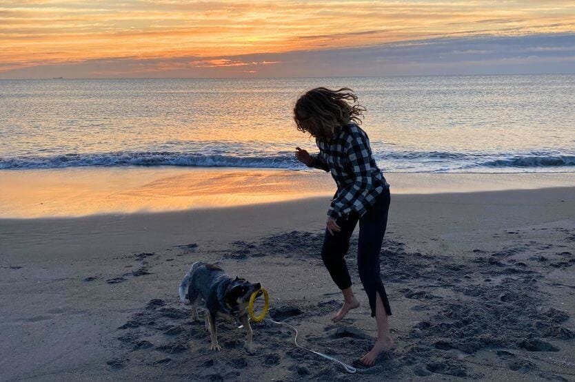 Scout, a blue heeler holding a yellow tug toy ring, and Haley, a young woman in a black and white flannel, play together on Cocoa Beach at sunrise