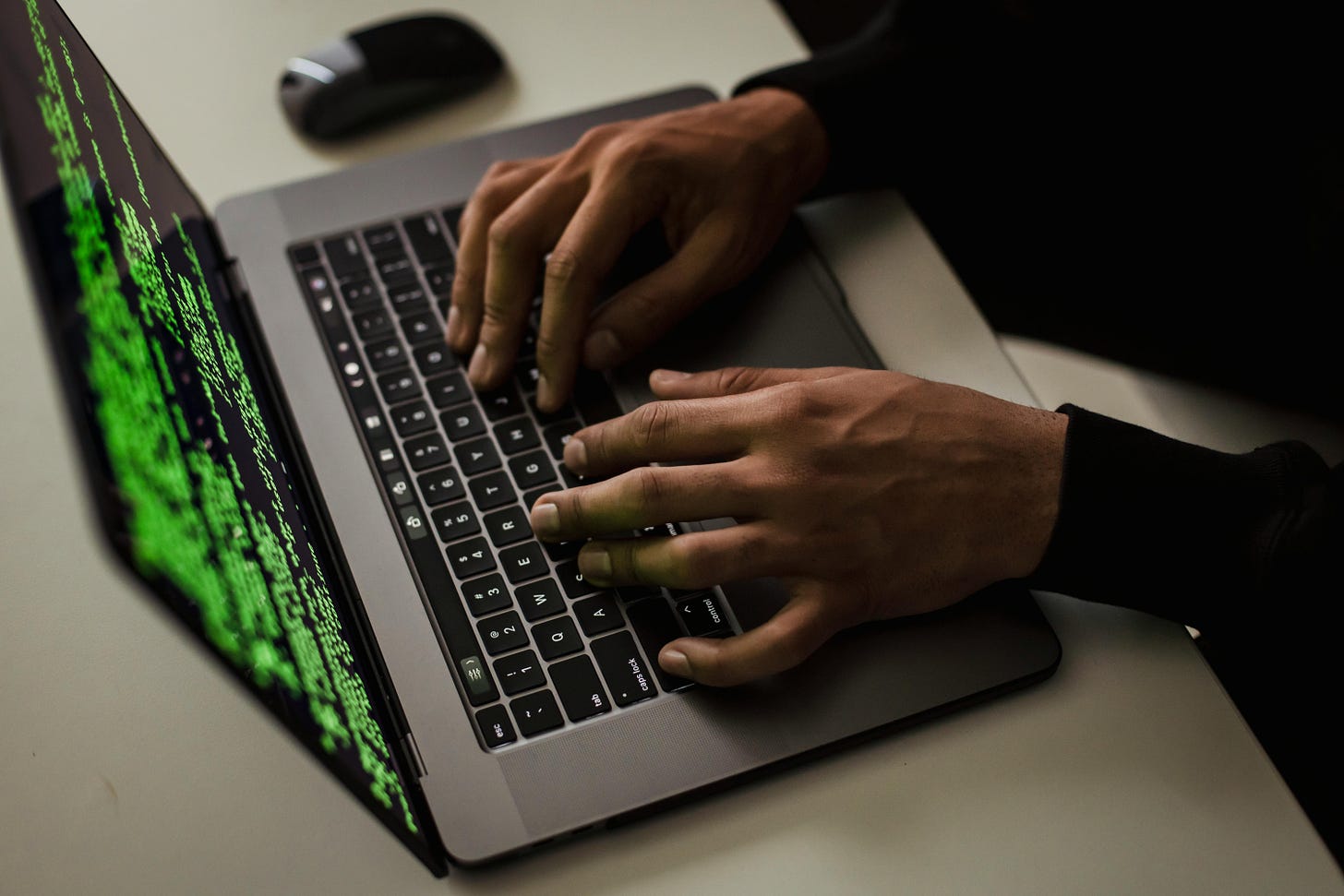 A photo taken from above of a silver coloured laptop with computer code on the screen. The code is bright green on a black background. A person's hands are visible resting on the keys.