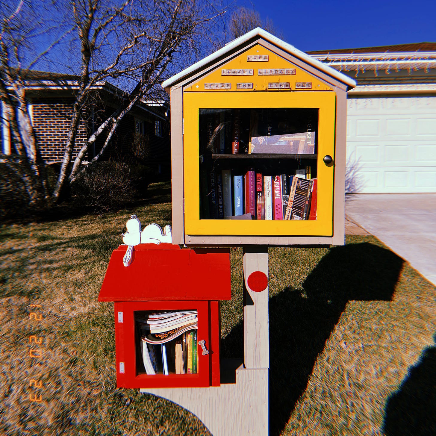 A photo of a little free library. It has two levels, one yellow and one red, with Snoopy lounging on the red one.