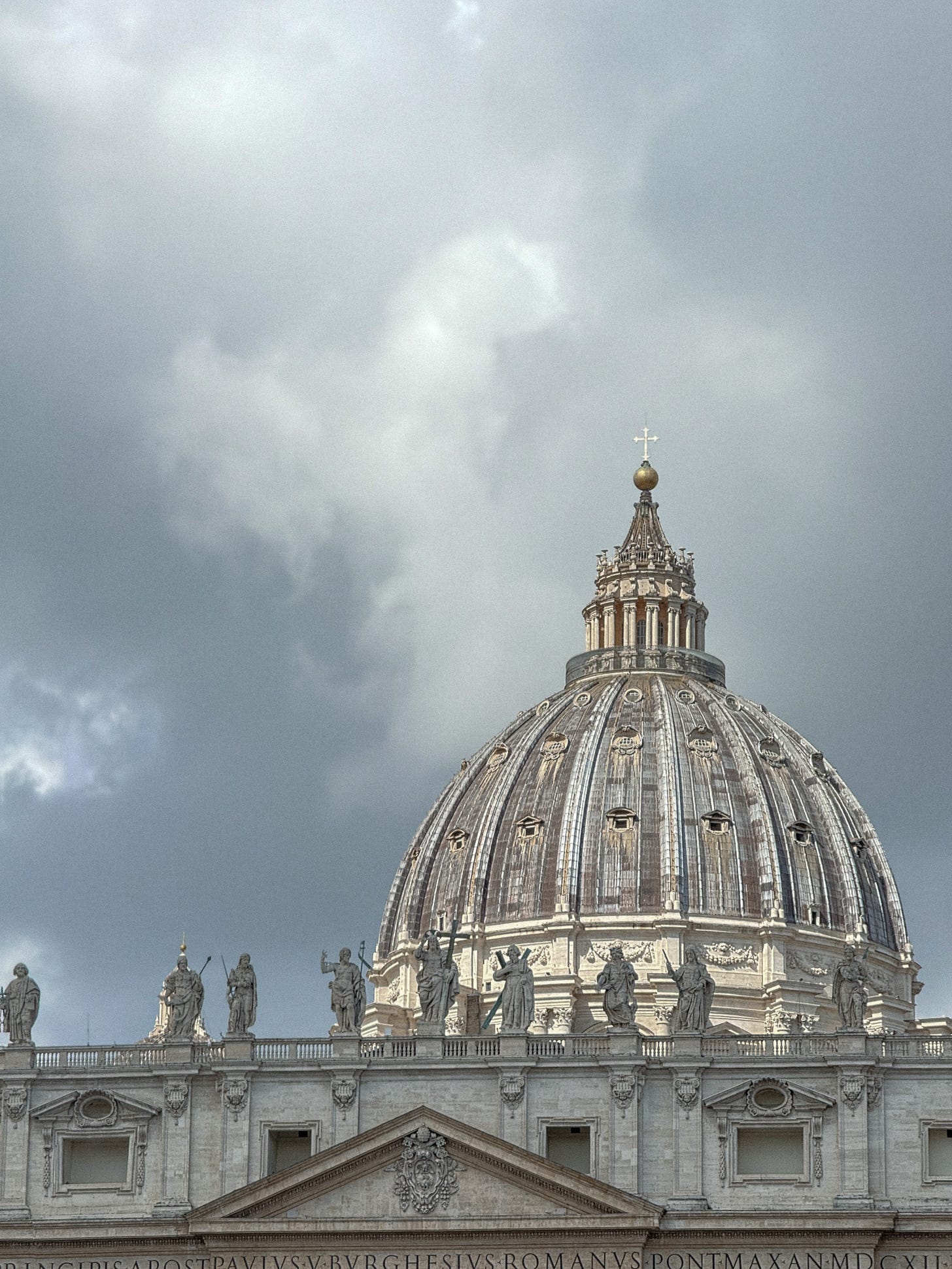 St. Peter’s Basilica dome rising against a muted, cloud-filled sky, detailed sculptures lining the rooftop