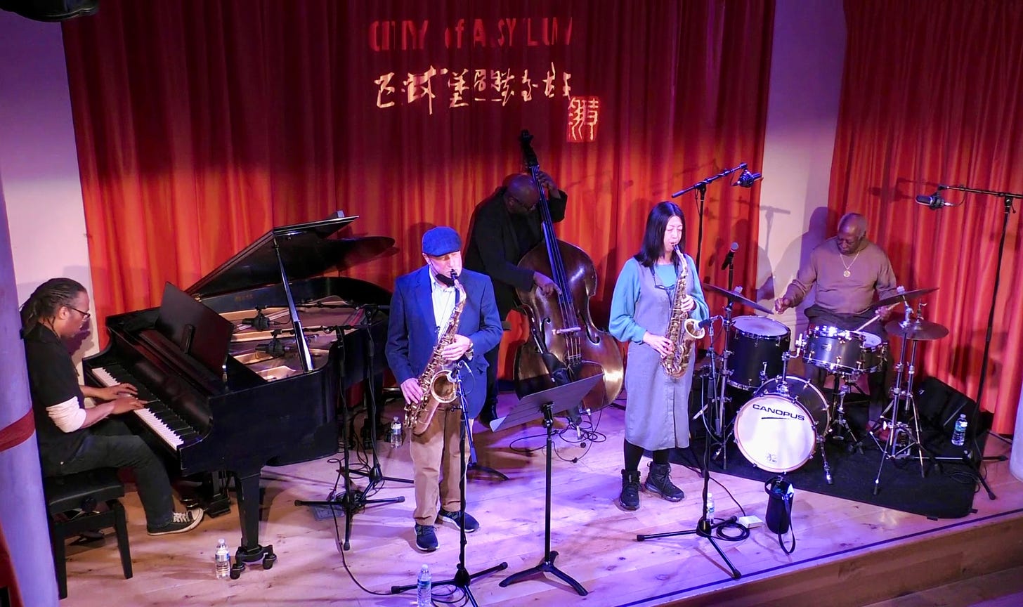 Image of a five-piece jazz band playing live. There is a drummer, two saxophone players (tenor and alto), a pianist, and an upright bass player.