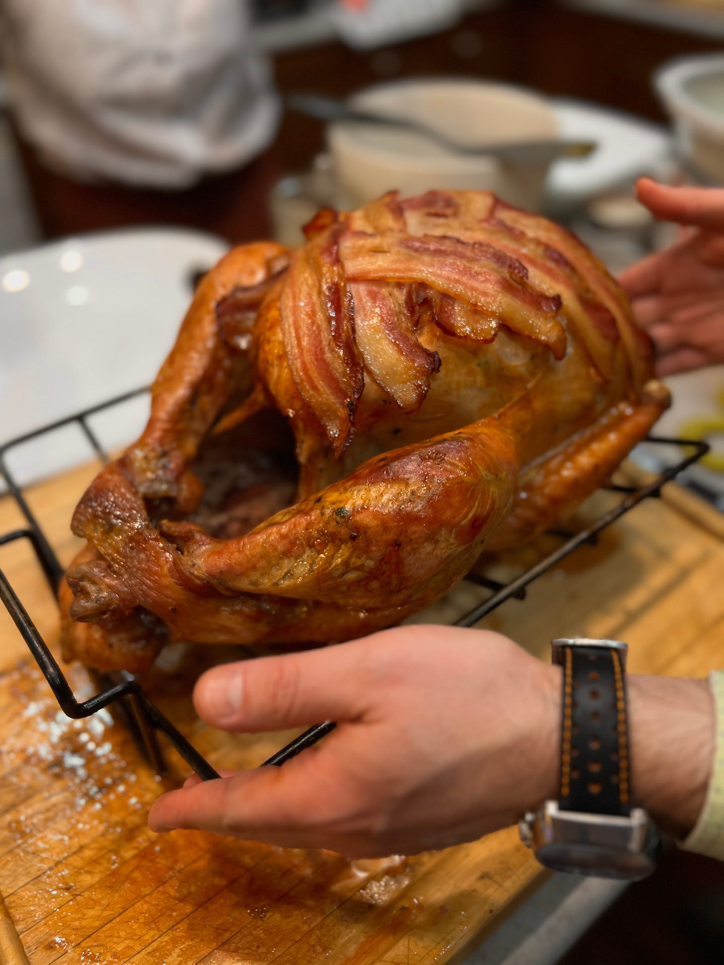 A turkey resting on a rack over a cutting board. Bacon slices are latticed across the top and everything is nicely brown. Jeff's hands hover at the bottom left and top right of frame, surrounding it.