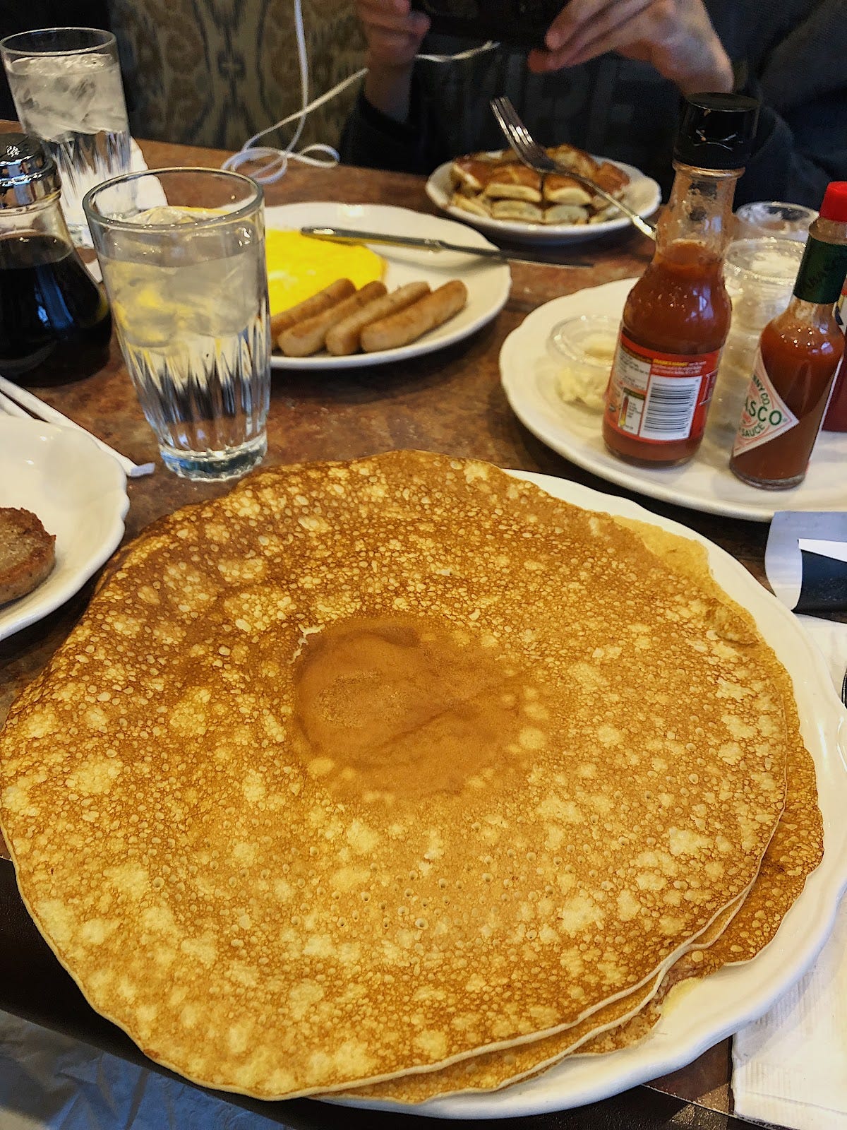 A stack of giant 49er pancakes and sausage links in the distance and sides of butter and tabasco sauce on plates at a breakfast restaurant