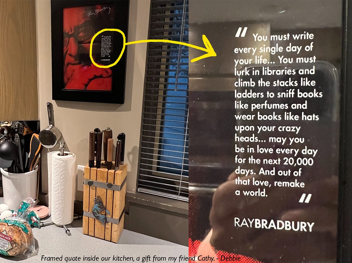 The image shows a kitchen countertop with a wooden knife block, paper towel holder, and various kitchen utensils. Above the countertop hangs a framed picture of Ray Bradbury, featuring a quote in white text. The quote reads: "You must write every single day of your life... You must lurk in libraries and climb the stacks like ladders to sniff books like perfumes and wear books like hats upon your crazy heads... may you be in love every day for the next 20,000 days. And out of that love, remake a world." The image includes an annotation with a yellow arrow pointing to the quote, emphasizing its importance. The bottom of the image is captioned, "Framed quote inside our kitchen, a gift from my friend Cathy. - Debbie."