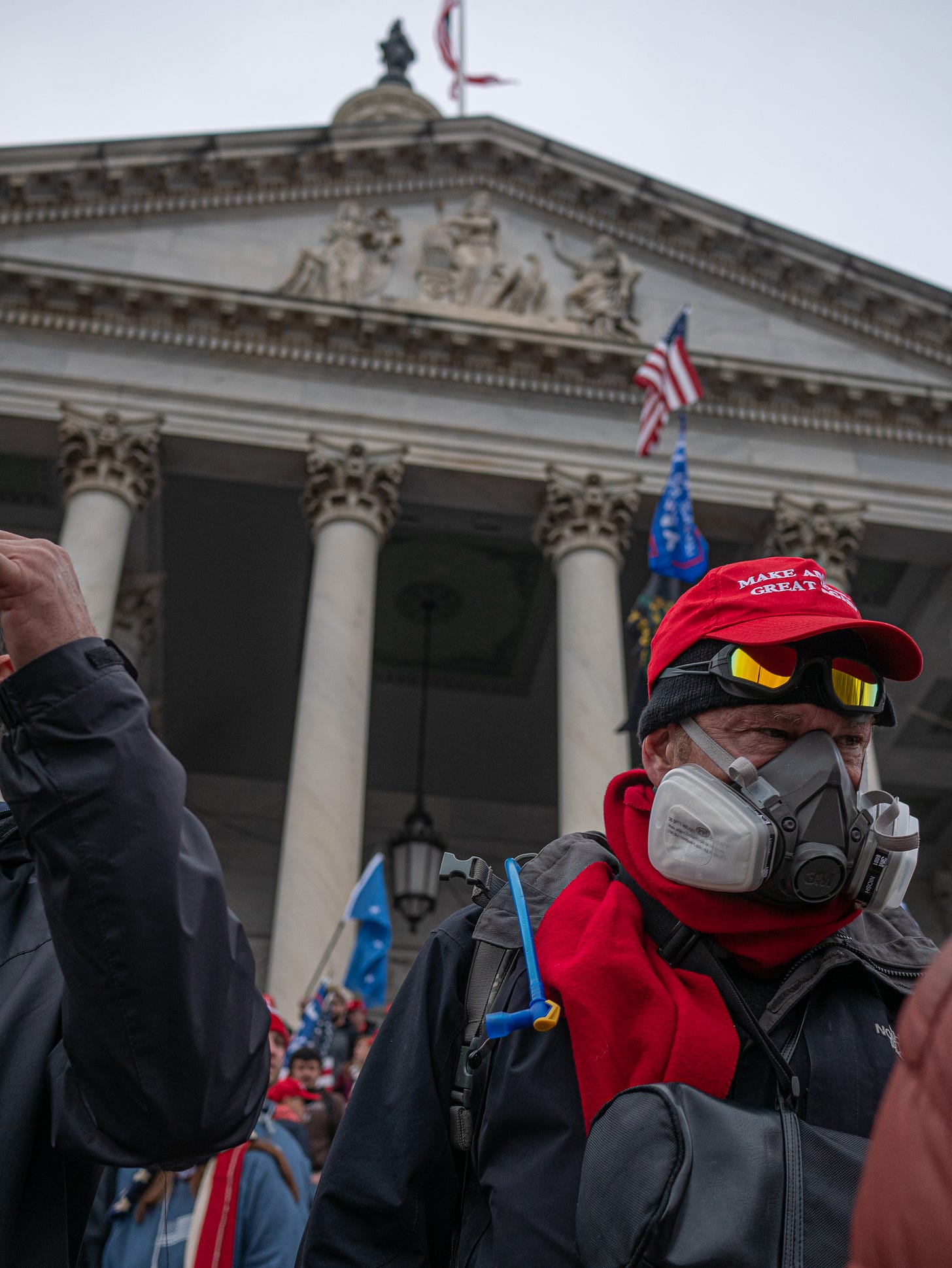 An older, middle-aged man wearing a red Make America Great Again hat over a fleece cap stares intently down the steps of the US Capitol, Jan 6, 2021. He is wearing a respiratory mask, the kind you might see at a construction site. He is wearing a red sweatshirt beneath a back jacket, and a backpack with the tube of a water reservoir slung over his right shoulder. Behind him are the arches and pediment of the US Capitol.