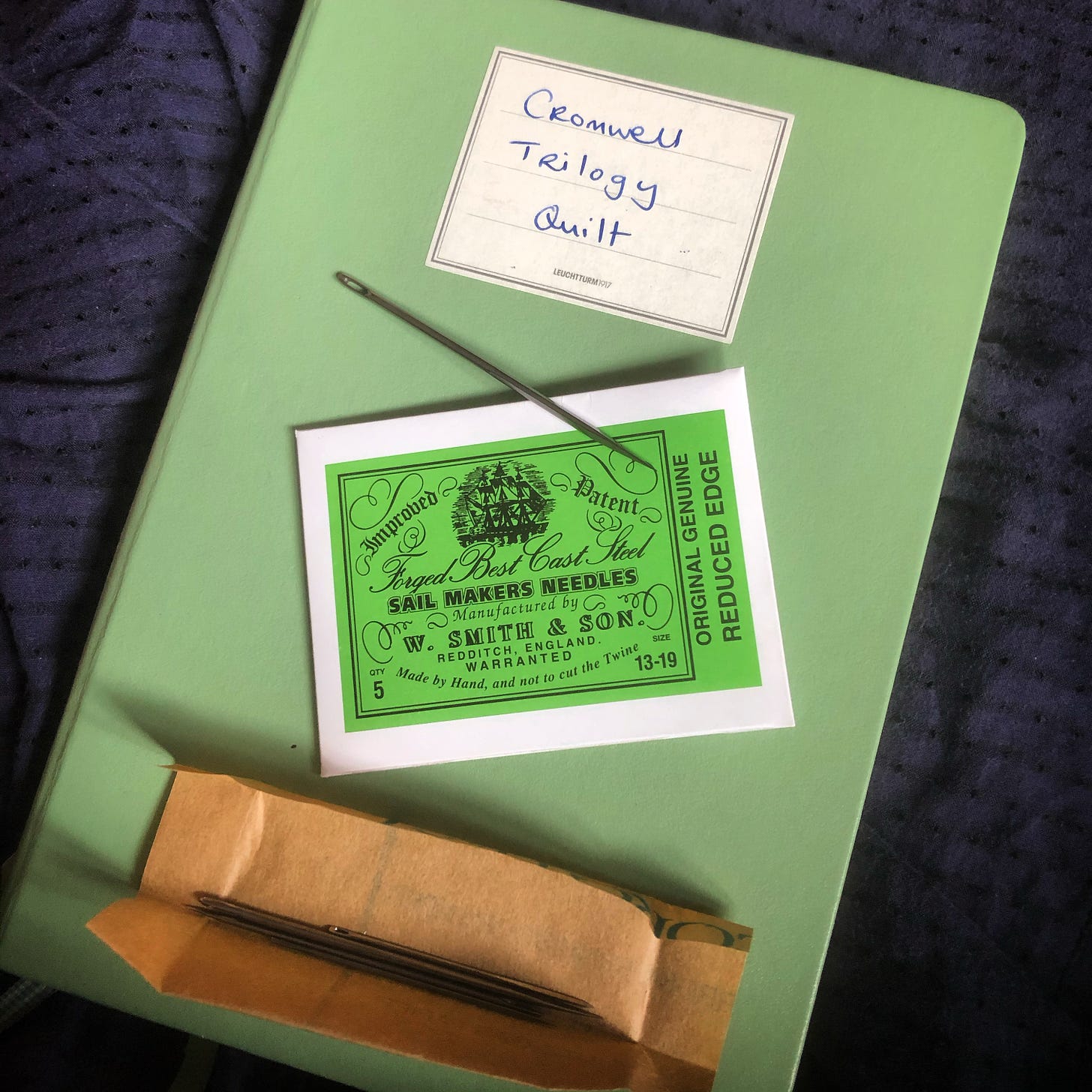 A green notebook labelled “Cromwell Trilogy Quilt”. On the top, there is a green packet marked “Sail Makers Needles”, and a large thick needle is visible.