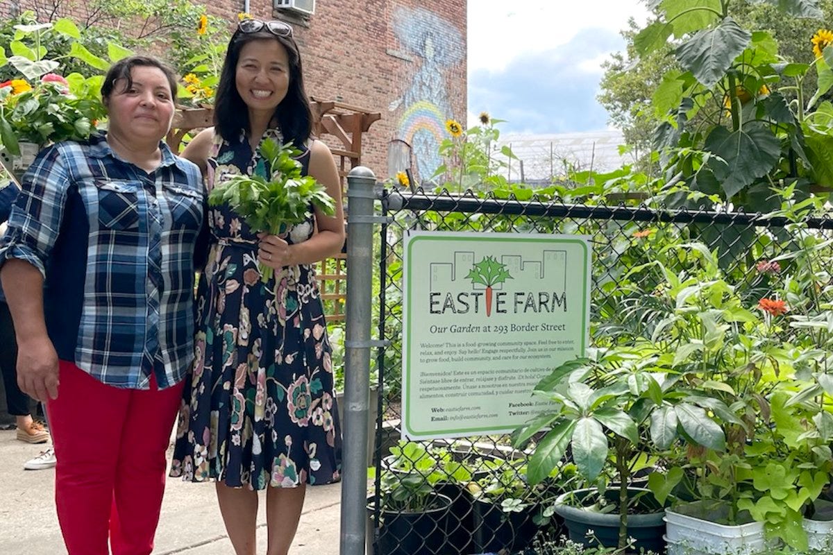 Michelle poses for a picture with a staff member of Eastie Farm while holding a bunch of greens and standing next to the Eastie Farm sign