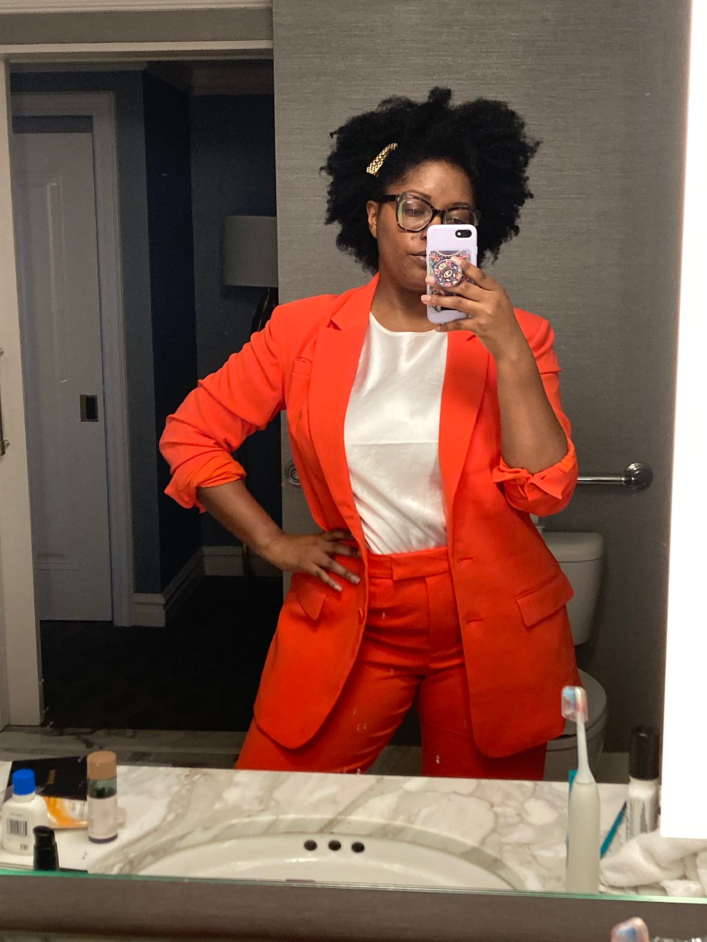 Black woman with a fro and glasses wearing a bright orange suit and satin white shirt taking a selfie in a bathroom mirror.