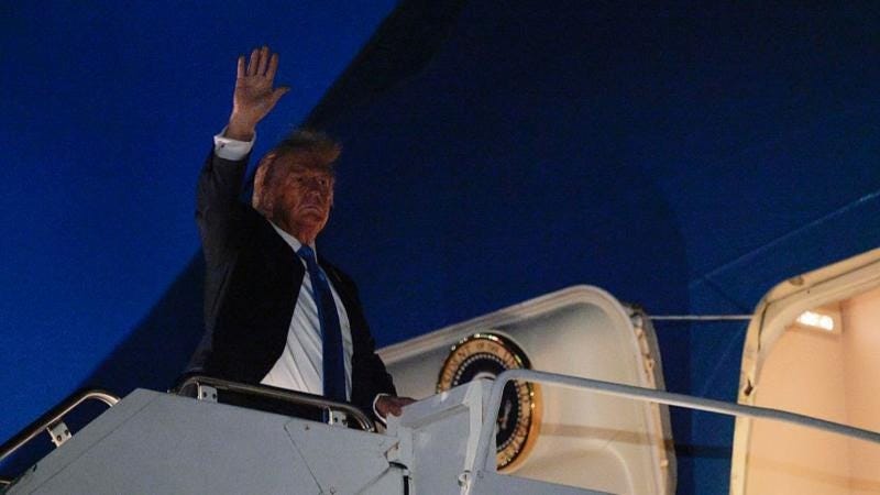 Trump wears a dark-coloured suit, white shirt an blue tie and stands at the top of a set of stairs to enter a plane. It is night time and dark and he is waving
