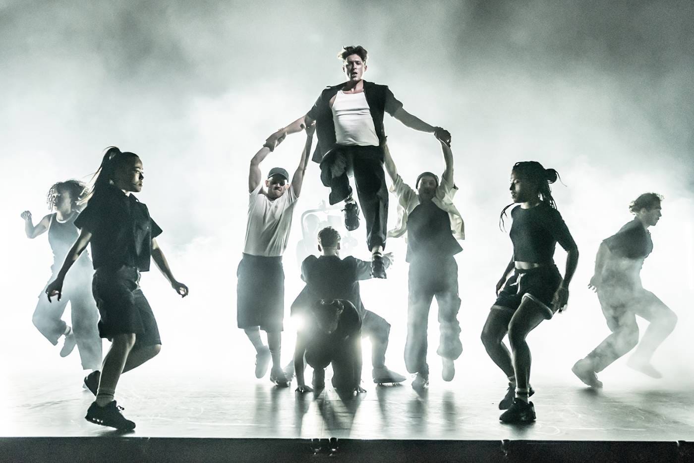 Tom Francis and the ensemble of Sunset Boulevard on the West End. Francis is being lifted up by a few dancers in a dramatic photo.