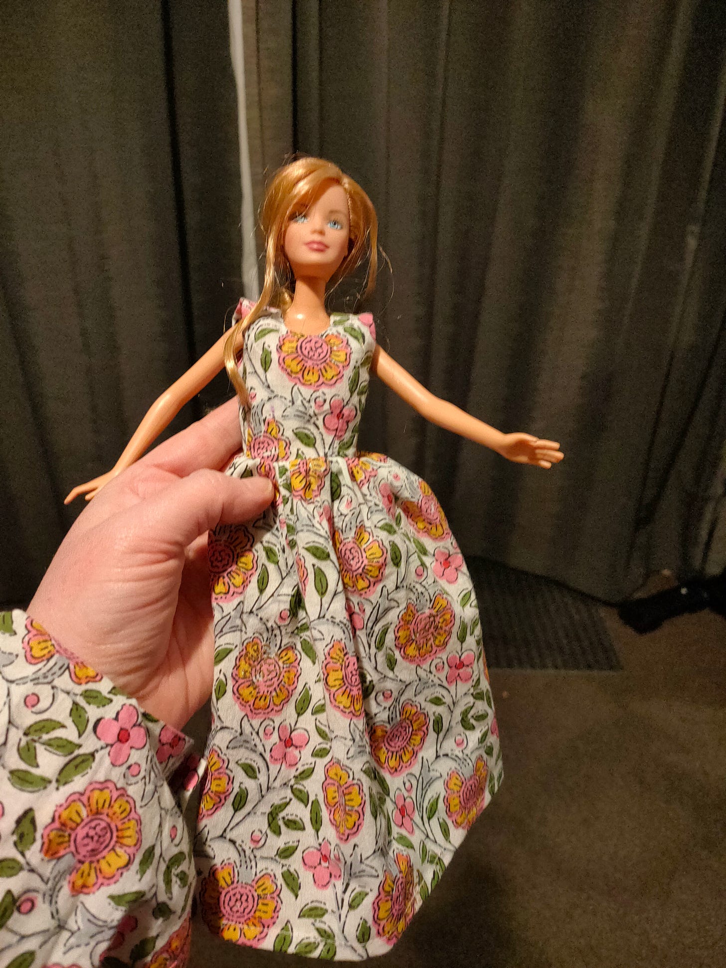 A fuzzy picture of a blonde barbie doll wearing a dress with a yellow, pink, green floral pattern long dress, with hints of gold glimmer. The hand holding the Barbie is showing a shirt cuff made from the same material. What is a midsize floral print on the human is an enormous STATEMENT PRINT on the Barbie.