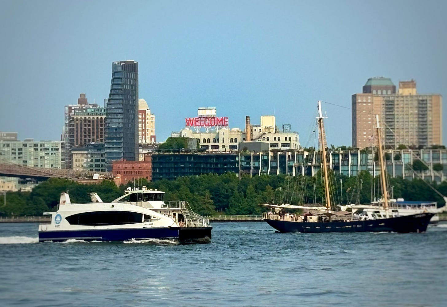 Looking back at DUMBO from the ferry to Manhattan. And old fashioned sailing ship and a city ferry begin to cross paths. A red WELCOME sign hangs over Brooklyn.