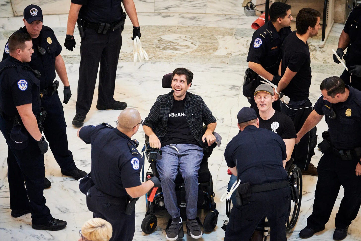 Mr. Barkan, photographed from above, is smiling as he sits in a wheelchair beside another protester in a wheelchair as police officers surround them. 