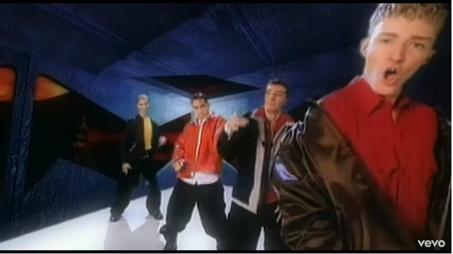 NSYNC has changed to button up tops and gigantic jackets, a different color for each guy. Pants are the same. They're dancing in a new spot now. For some reason this screenshot makes it look like Justin Timberlake has only two teeth.