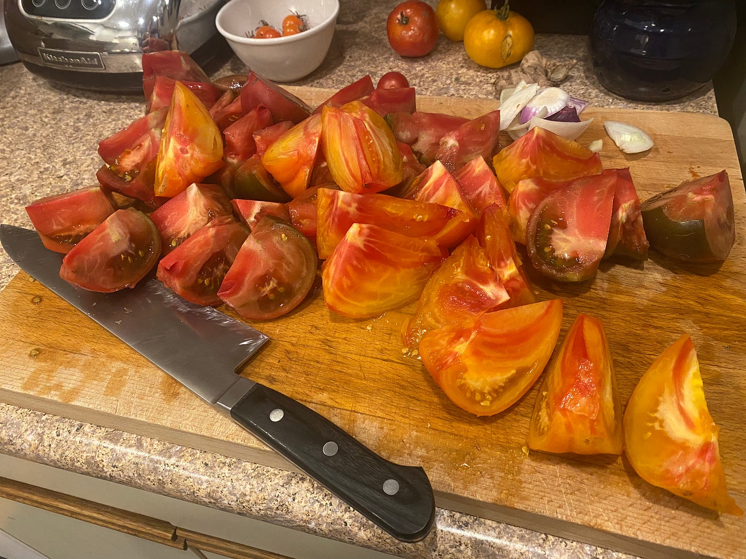 A wooden cutting board full of heirloom tomatoes cut into thick wedges. The knife is tucked under a few of them at the bottom left corner. In the upper right corner are scraps of onion.