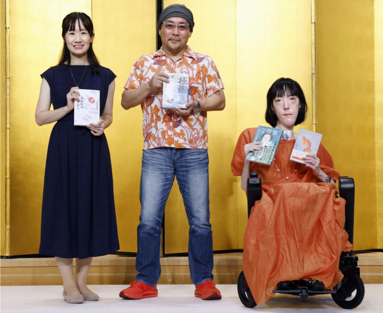 3 award winners hold their books, including a disabled woman in a long flowing orange dress in a black powerchair.