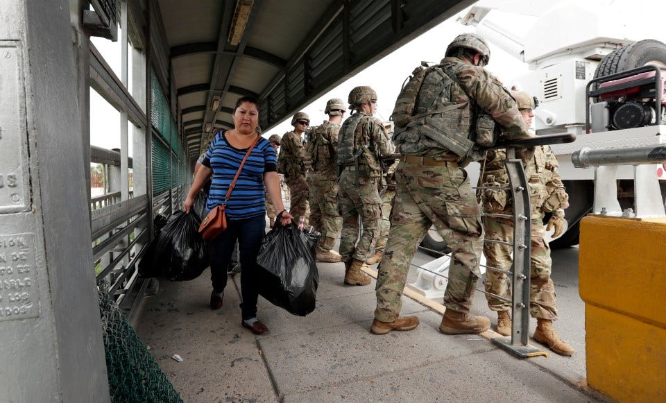 A woman carrying bags walks past U.S. military personnel installing razor wire on a border bridge.