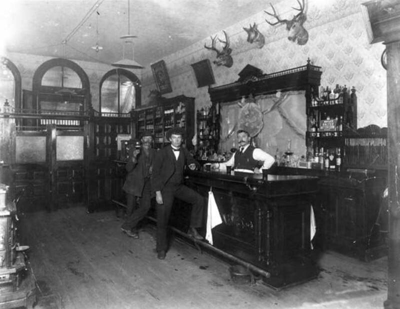 A black and white photo of a vintage bar. Two men are standing in front of the bar counter, while a bartender serves drinks behind it. The bar is decorated with mounted deer heads and various bottles. The room has high ceilings, and ornate woodwork is visible throughout.