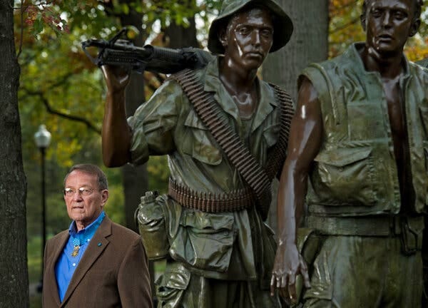 Roger Donlon in front of a statue of two weary-looking servicemen, one of whom holds a machine gun on his shoulder.