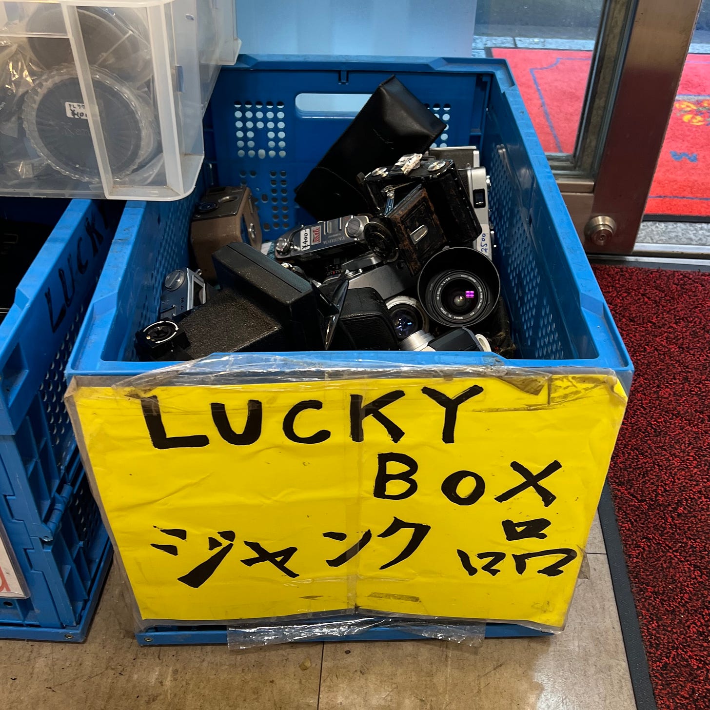 Blue plastic crate full of vintage cameras with bright yellow sign reading in English "LUCKY BOX", and in Japanese "Junk"