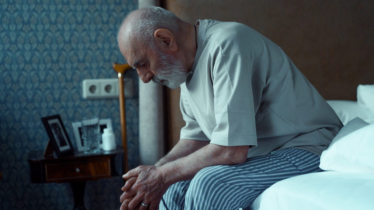 An old man sits on the bed looking at his hands in a depressed manner