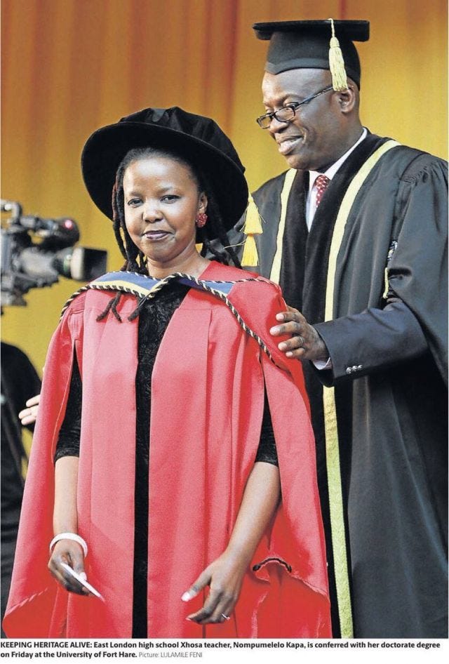 A South African woman dressed in a red gown and black velvet cap, with a South African man in a black academic robe standing behind her