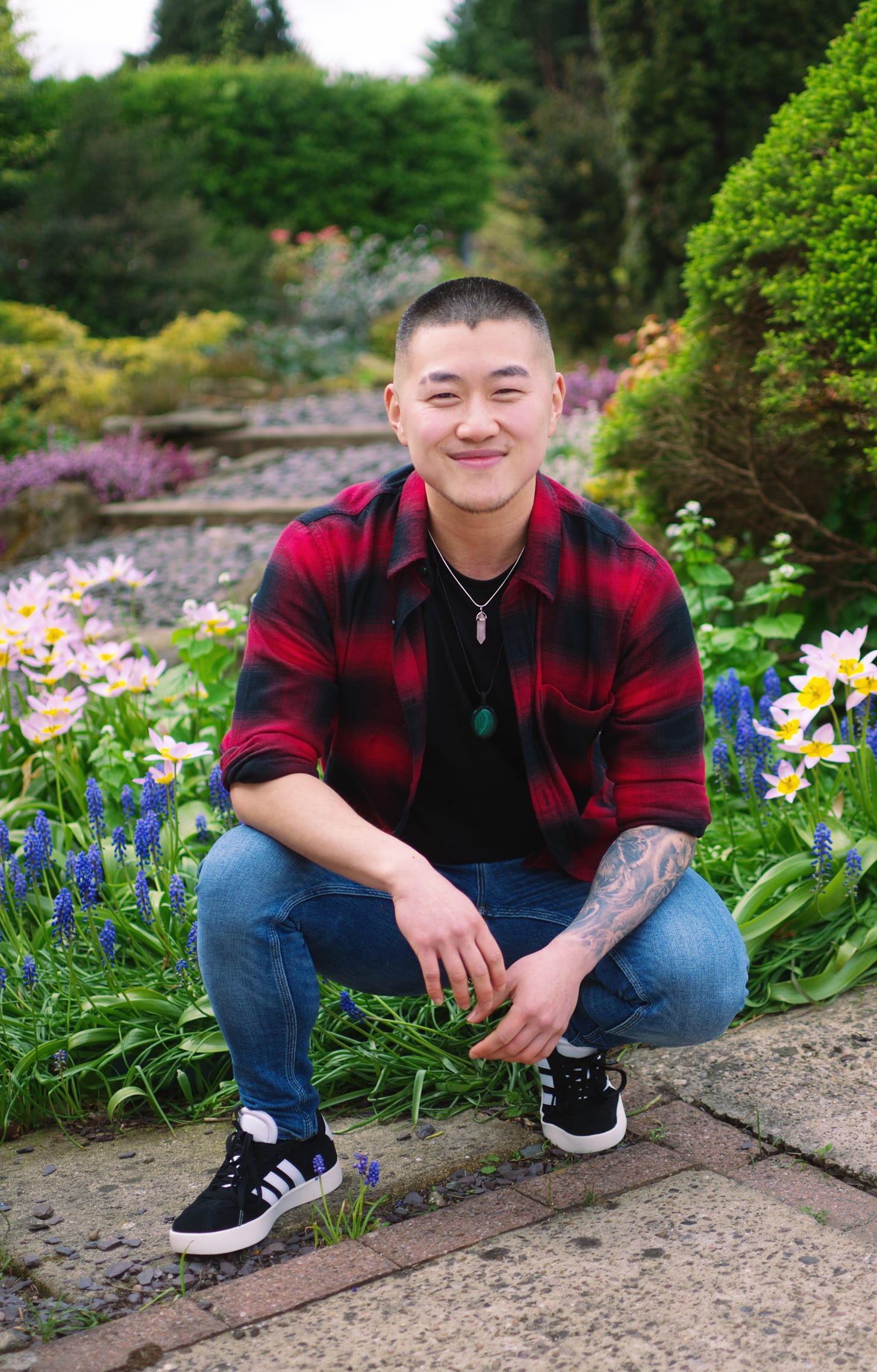 A Chinese person wearing a red and black flannel shirt and blue jeans crouches in front of a small bed of purple flowers. They have a shaved head and are smiling, looking directly at the camera.