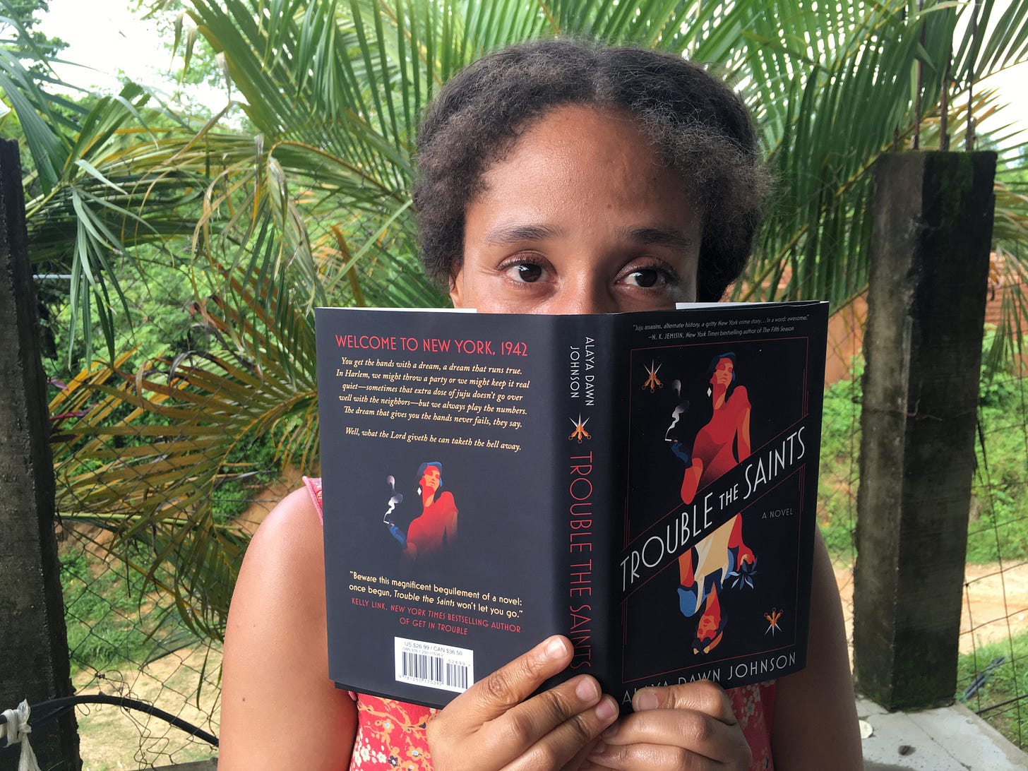 A woman hides most of her face behind a novel called "Trouble the Saints" with palm fronds and a dirt road in the background.