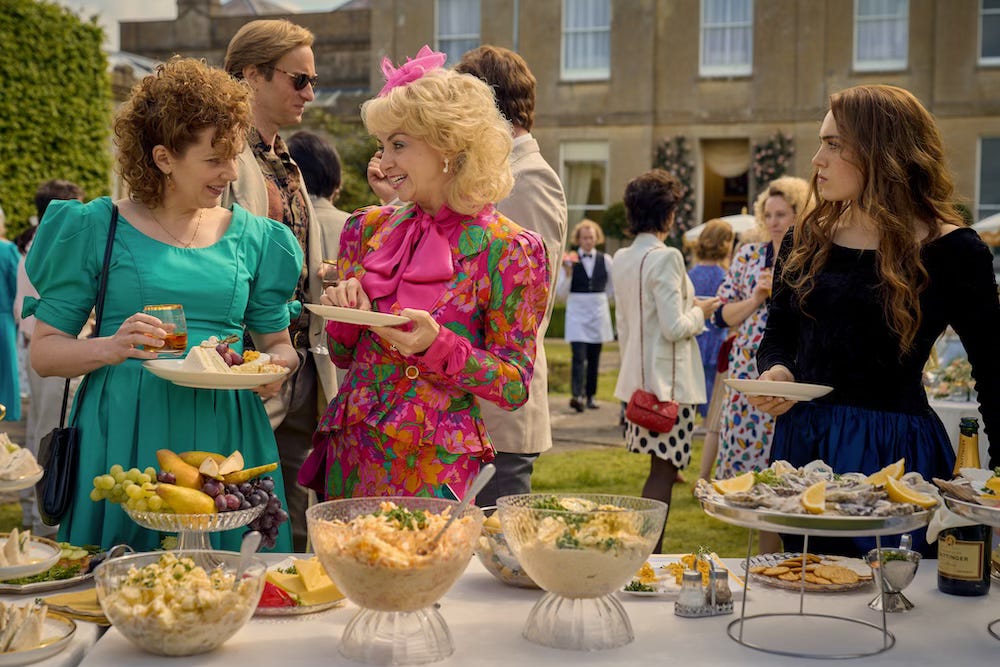 Katherine Parkinson, Lisa McGrillis, and Bella Maclean in a party scene from Rivals, standing near a table of food