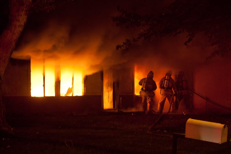 A house is consumed with fire and several firefighters are at the front door with a hose and other equipment.