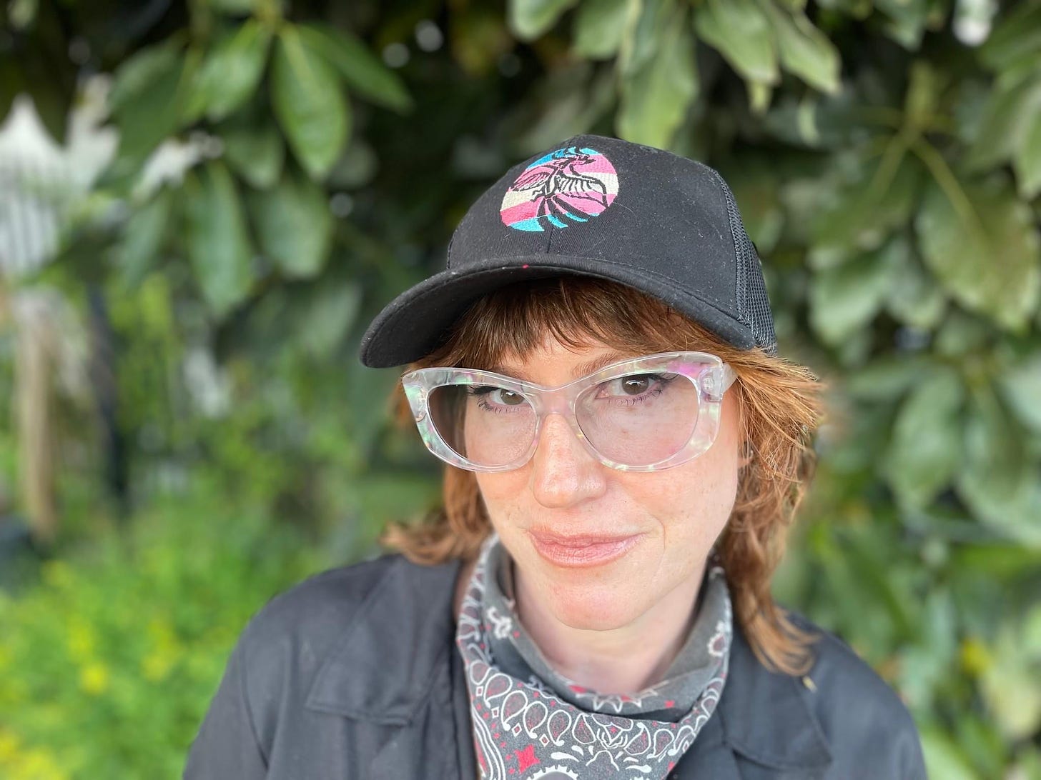 Lasara, a genderqueer white person with red hair and glasses, wearing black coveralls, a cap, and a bandana, looks at the camera.