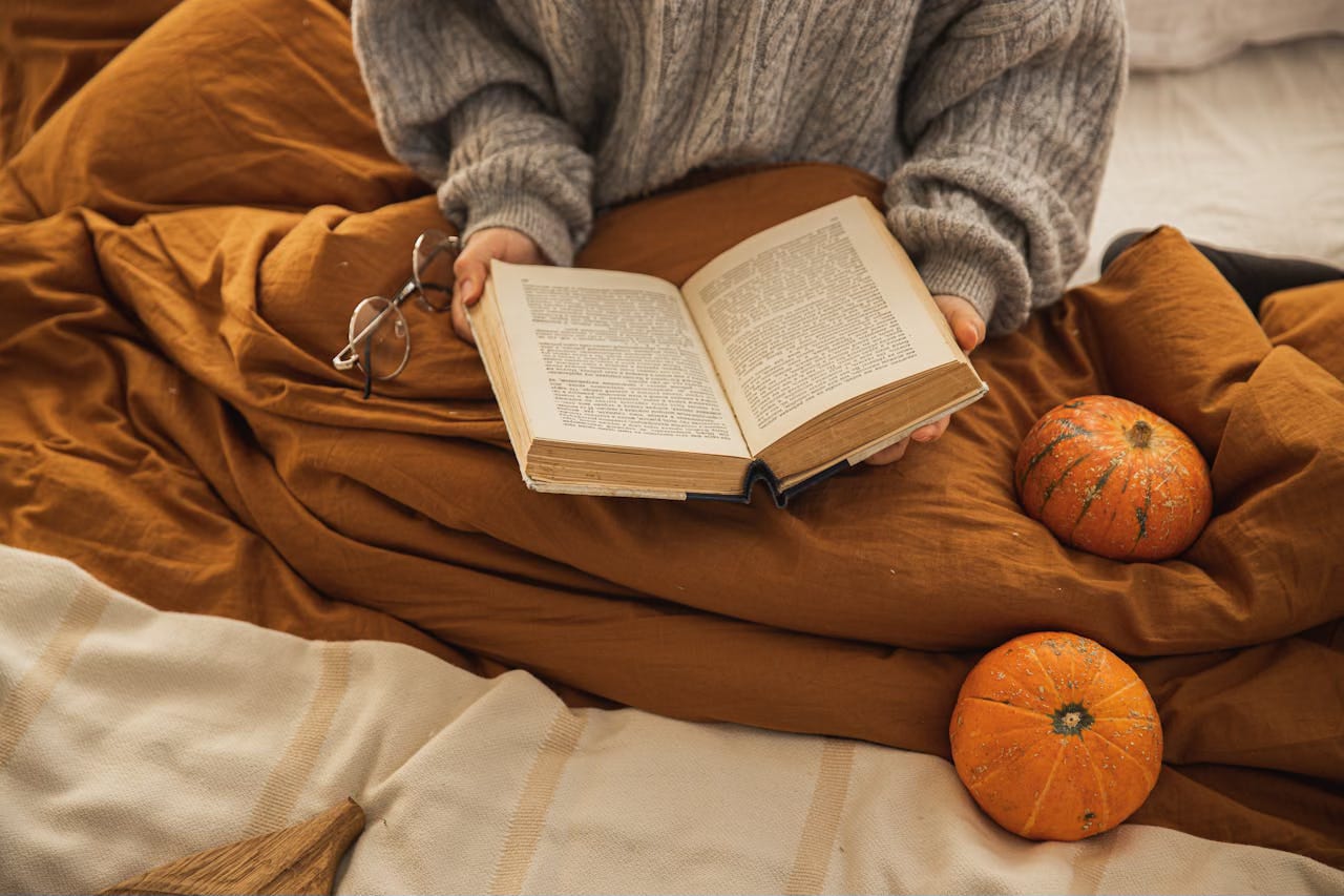 Woman sitting on the floor wrapped in a dark orange blanket reading a book with pumpkins next to her.