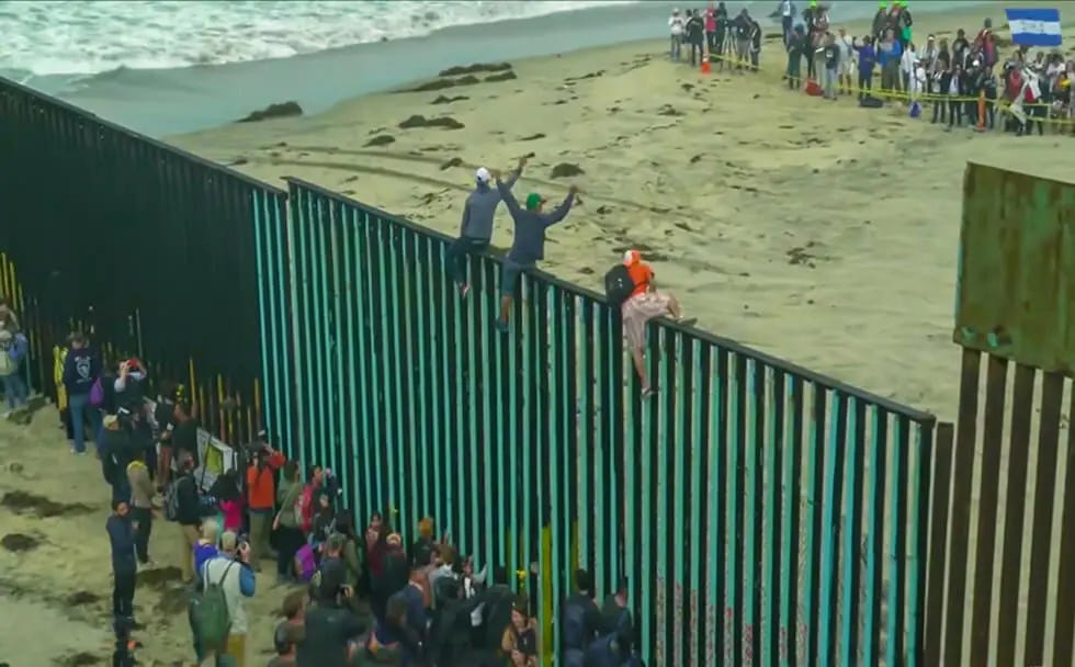 People gathered at a tall border fence by the beach, with some climbing on it