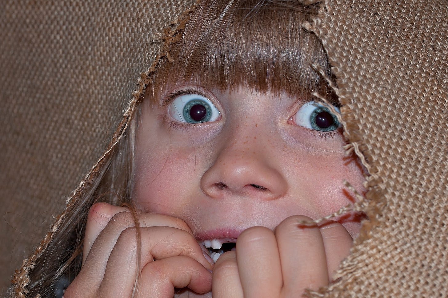 Frightened child with big blue eyes looking through a ripped brown cloth