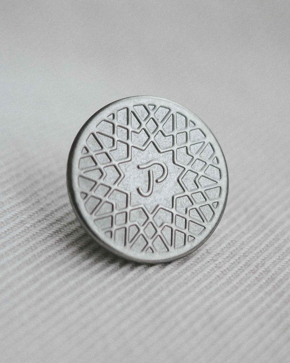 A close up photograph of the manhole-inspired button for Paynter's jacket. The button is a silver-coloured metal with an embossed letter P at the centre, surrounded by a series of star-shaped lines.