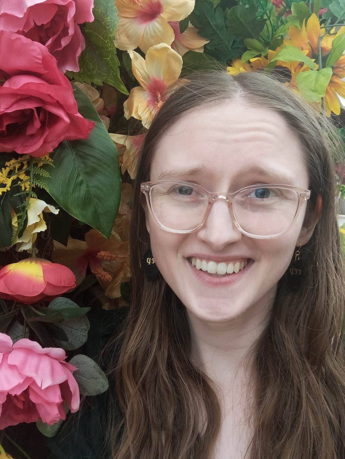 A selfie of Kandi Zeller (she/her), a white woman with long brown hair and pink glasses, with a backdrop of silk flowers that are hanging up in a craft store aisle