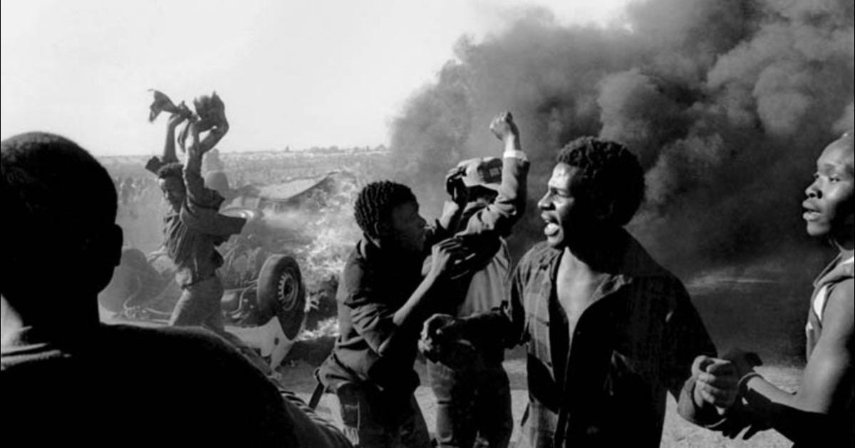 Black-and-white photo of a scene of chaos, Black bodies in shadow moving in the fore and midground while smoke and upturned vehicles are present in the background of a fraught anti-apartheid protest in the 1980s.