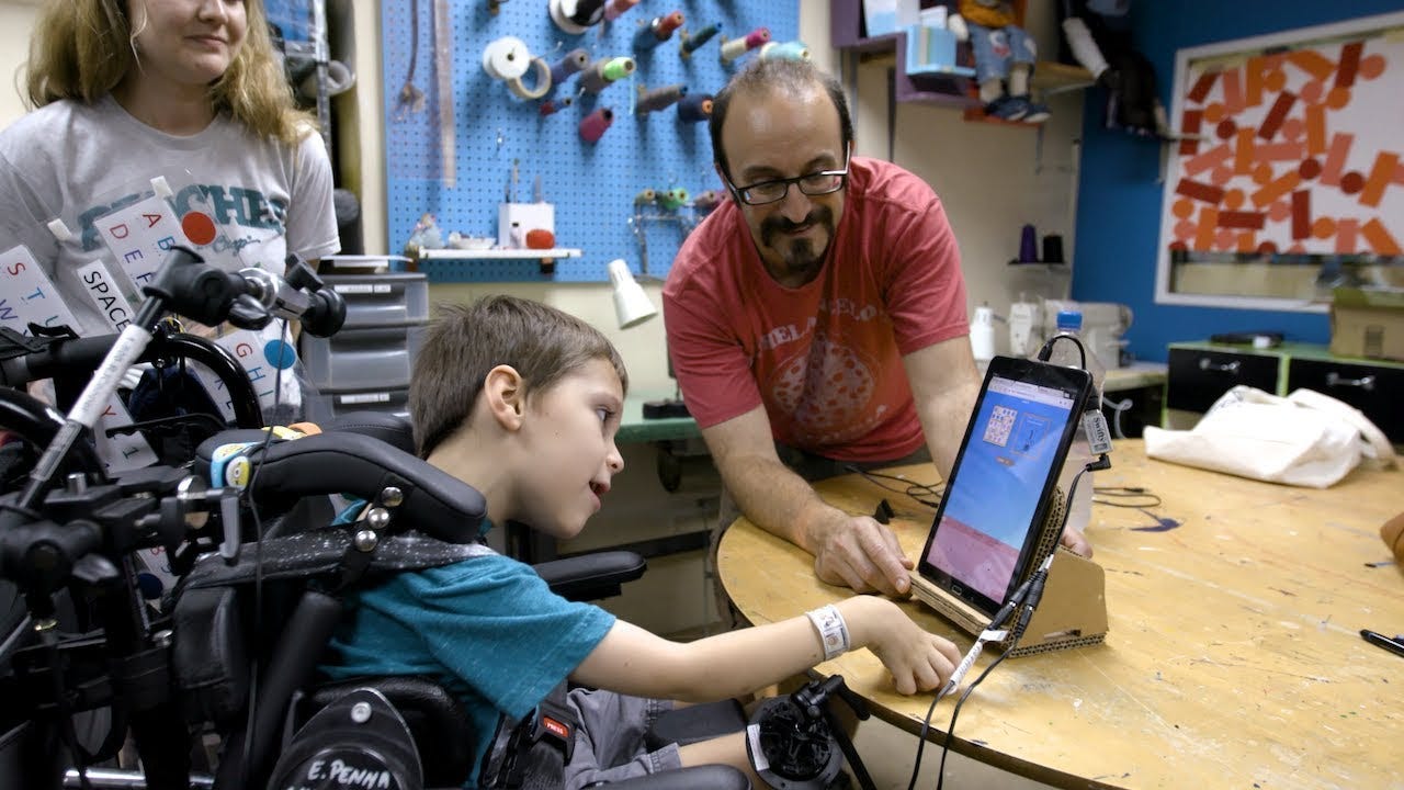 A child in a motorized wheelchair lifts his hand to interact with a digital tablet on a table, with customized tablet support made from cardboard by the Adaptive Design Association. A parent and fabricator look on.