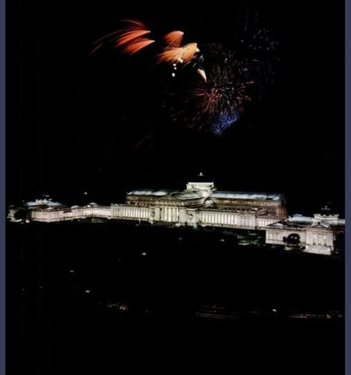 Chicago's Museum of Science and Industry illuminated against pitch black night sky, overhead orange and blue fireworks explode.